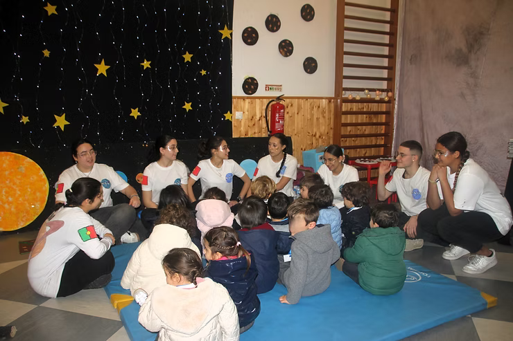 Grupo sentado em colchonetes azuis em uma sala de aula, conversando com os professores perto de uma parede preta estrelada e uma escada de madeira.