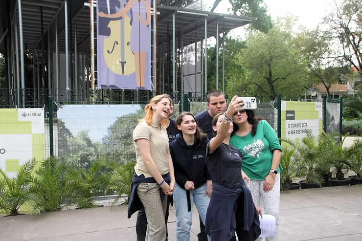 Quatro pessoas posando para uma selfie ao ar livre perto de um prédio com vegetação e uma placa grande.