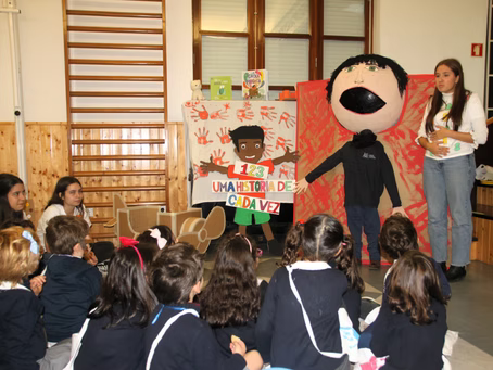 Crianças assistem a um fantoche e a um orador em uma sala de aula, com um cartaz colorido exposto na frente.