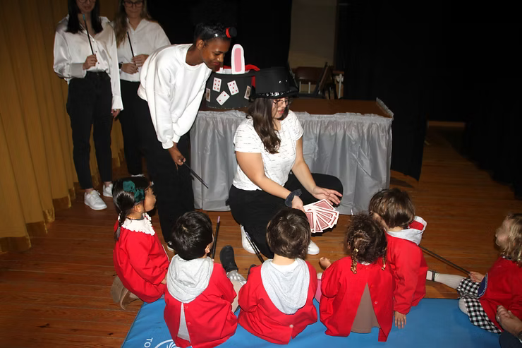 Crianças vestidas de vermelho sentam-se num tapete azul enquanto dois adultos apresentam um teatro de fantoches num palco de madeira.