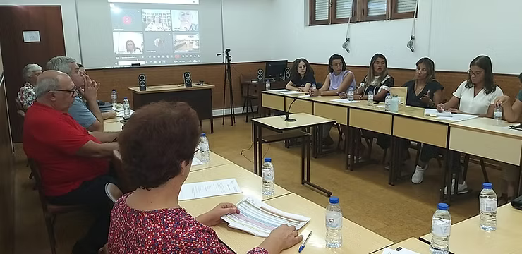 Sala de reuniões com pessoas sentadas ao redor de mesas, assistindo a uma apresentação projetada na parede.