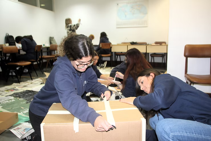 Pessoas em uma sala de aula montando uma caixa de papelão no chão.