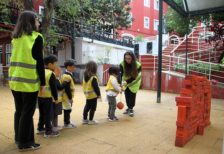 Crianças com coletes amarelos brincam de empilhar blocos ao ar livre, com dois adultos por perto.