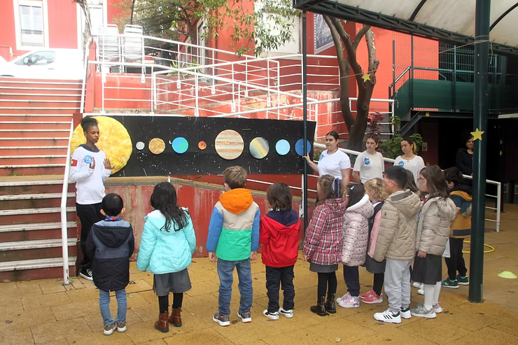 Crianças observam, em um parquinho, uma exposição com temática espacial que apresenta planetas em uma parede.
