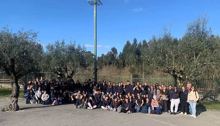 Um grande grupo posando ao ar livre em um parque ensolarado, com árvores, um poste de luz e uma montanha ao fundo.