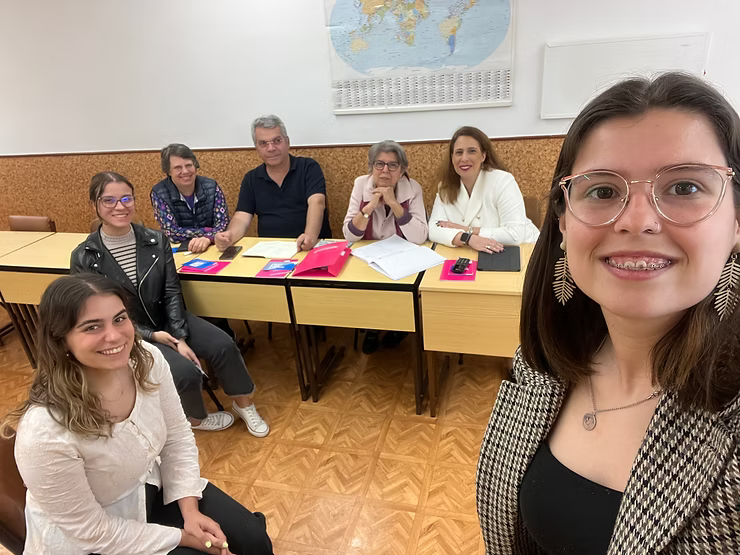 Grupo de pessoas sentadas em uma sala de aula, sorrindo ao redor de carteiras com cadernos e papéis.