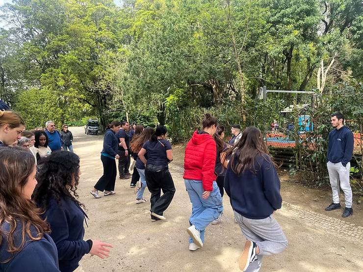 Pessoas dançando em uma clareira arborizada ao ar livre, com espectadores por perto.