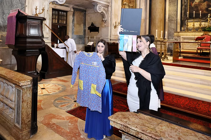 Três mulheres estão em um corredor da igreja examinando um tecido azul e amarelo perto de um púlpito de madeira.