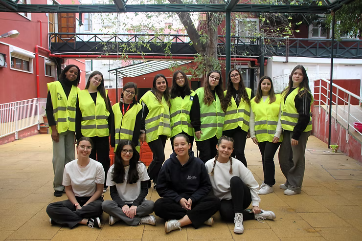 Grupo de pessoas posando em ambiente interno, vestindo coletes de segurança amarelo neon e roupas casuais, em uma passarela coberta.