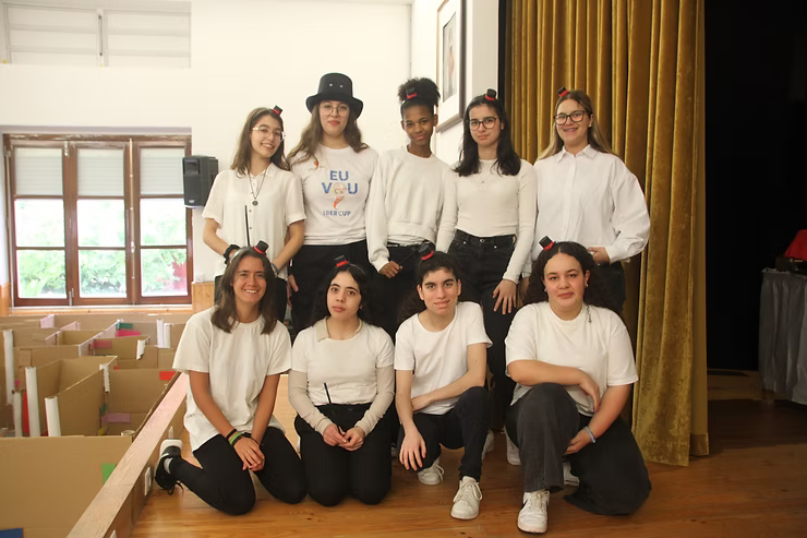 Grupo de oito pessoas posando em um palco dentro de casa, vestindo blusas brancas e calças pretas.