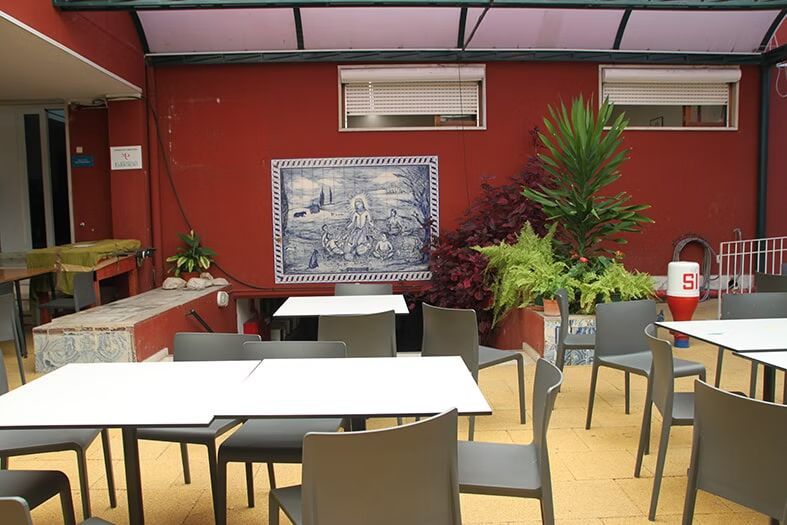 Pátio externo do café com mesas brancas, cadeiras bege e uma parede vermelha com um mural de azulejos e vasos de plantas.