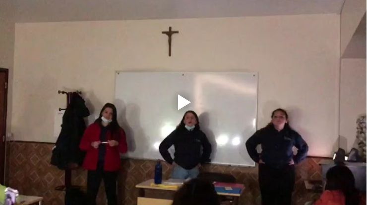 Três pessoas estão de pé em frente a um quadro branco sob uma cruz, em uma sala de aula ou sala de reuniões.