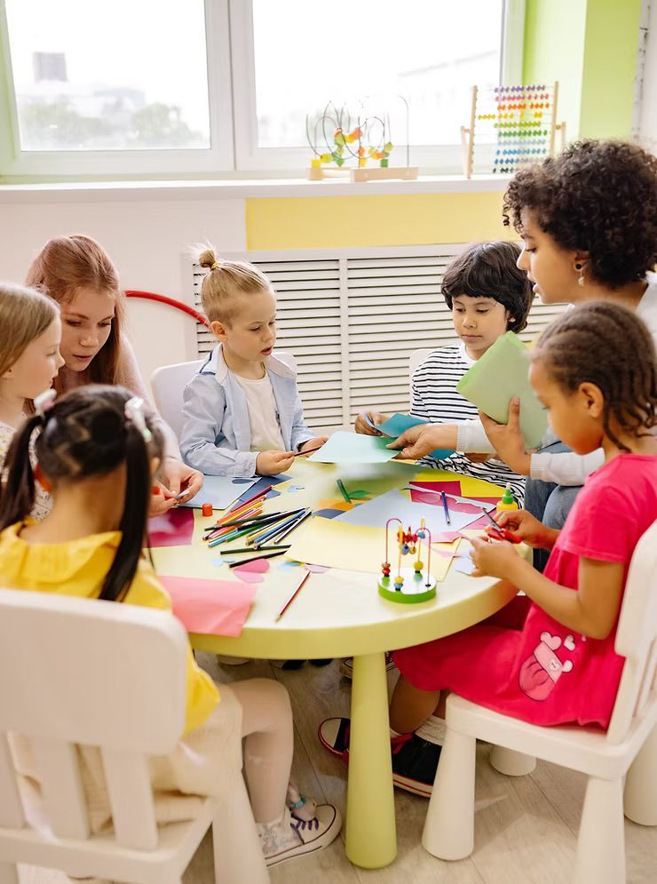 Crianças reunidas em volta de uma mesa, colorindo e fazendo trabalhos manuais em uma sala de aula iluminada.