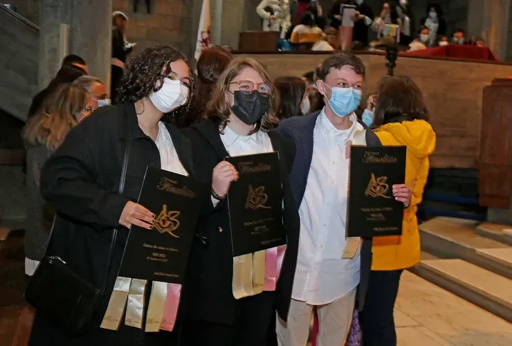 Três pessoas mascaradas seguram certificados pretos em uma cerimônia realizada em local fechado.
