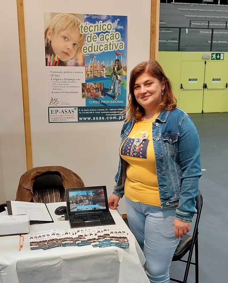 Mulher em pé ao lado de uma mesa de exposição em uma feira de educação tecnológica, com pôsteres e folhetos ao fundo.