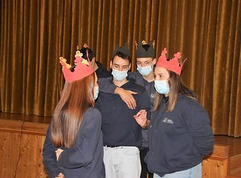 Três estudantes mascarados usando coroas de papel estão em um palco com uma cortina dourada ao fundo.
