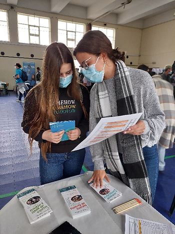Duas pessoas usando máscaras examinam folhetos em uma mesa durante um evento em local fechado.