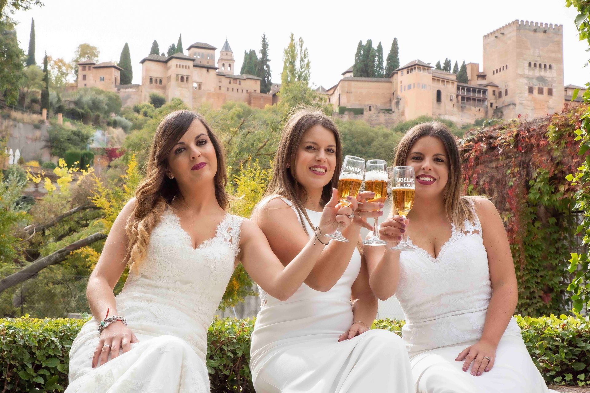 Tres mujeres con vestidos blancos están sentadas una al lado de la otra brindando con vasos de cerveza.