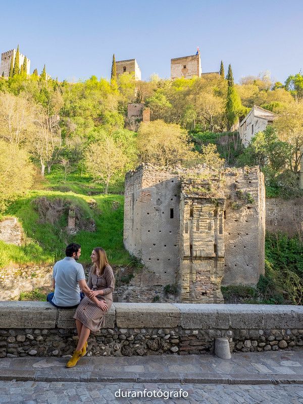 Un hombre y una mujer están sentados en un muro de piedra frente a un castillo.