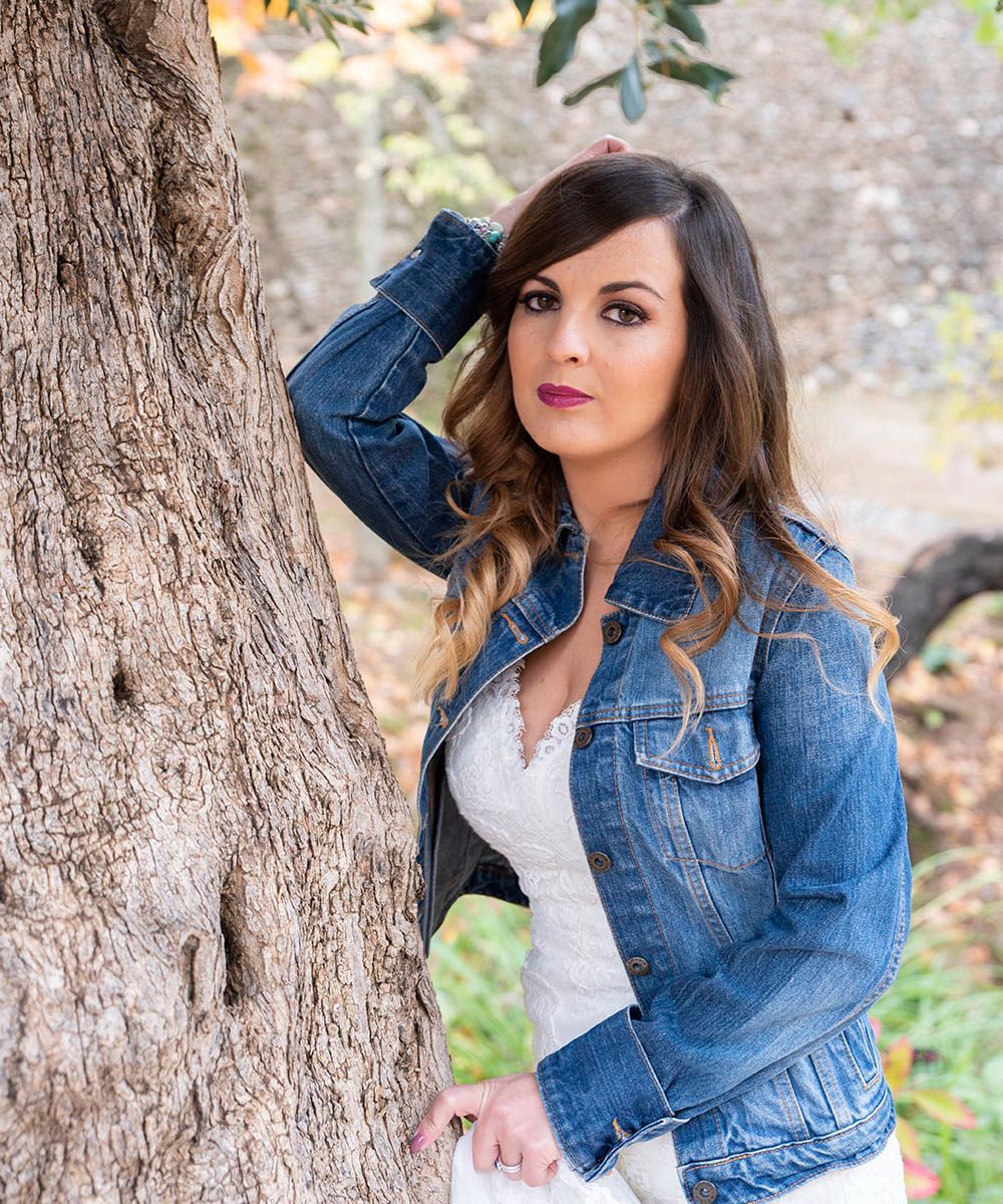 Una mujer con un vestido blanco y una chaqueta vaquera está apoyada contra un árbol.