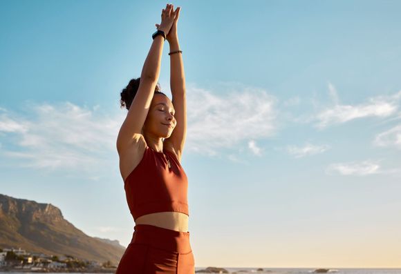 Mujer con ropa deportiva roja levanta los brazos al cielo, con los ojos cerrados. Vista al mar, día soleado.