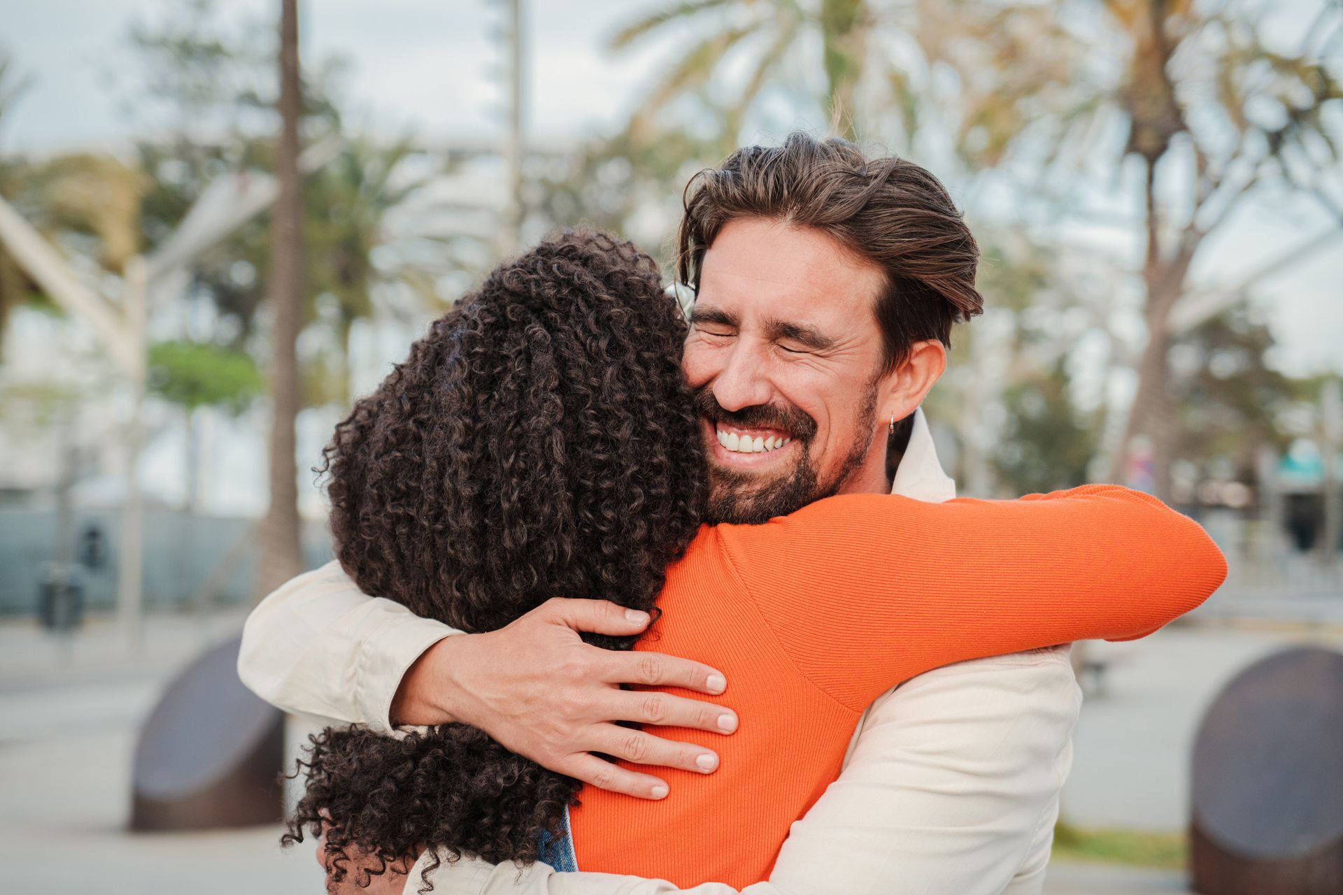 Hombre abraza a persona con camisa naranja, sonriendo ampliamente. Al aire libre, con palmeras al fondo.