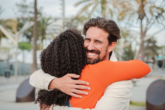 Hombre abraza a persona con camisa naranja, sonriendo ampliamente. Al aire libre, con palmeras al fondo.