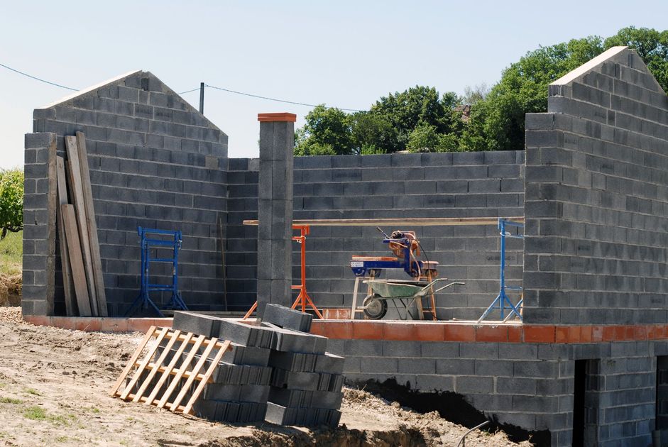 Maison en parpaings partiellement construite, avec des ouvriers sur des échafaudages et des matériaux de construction à proximité.