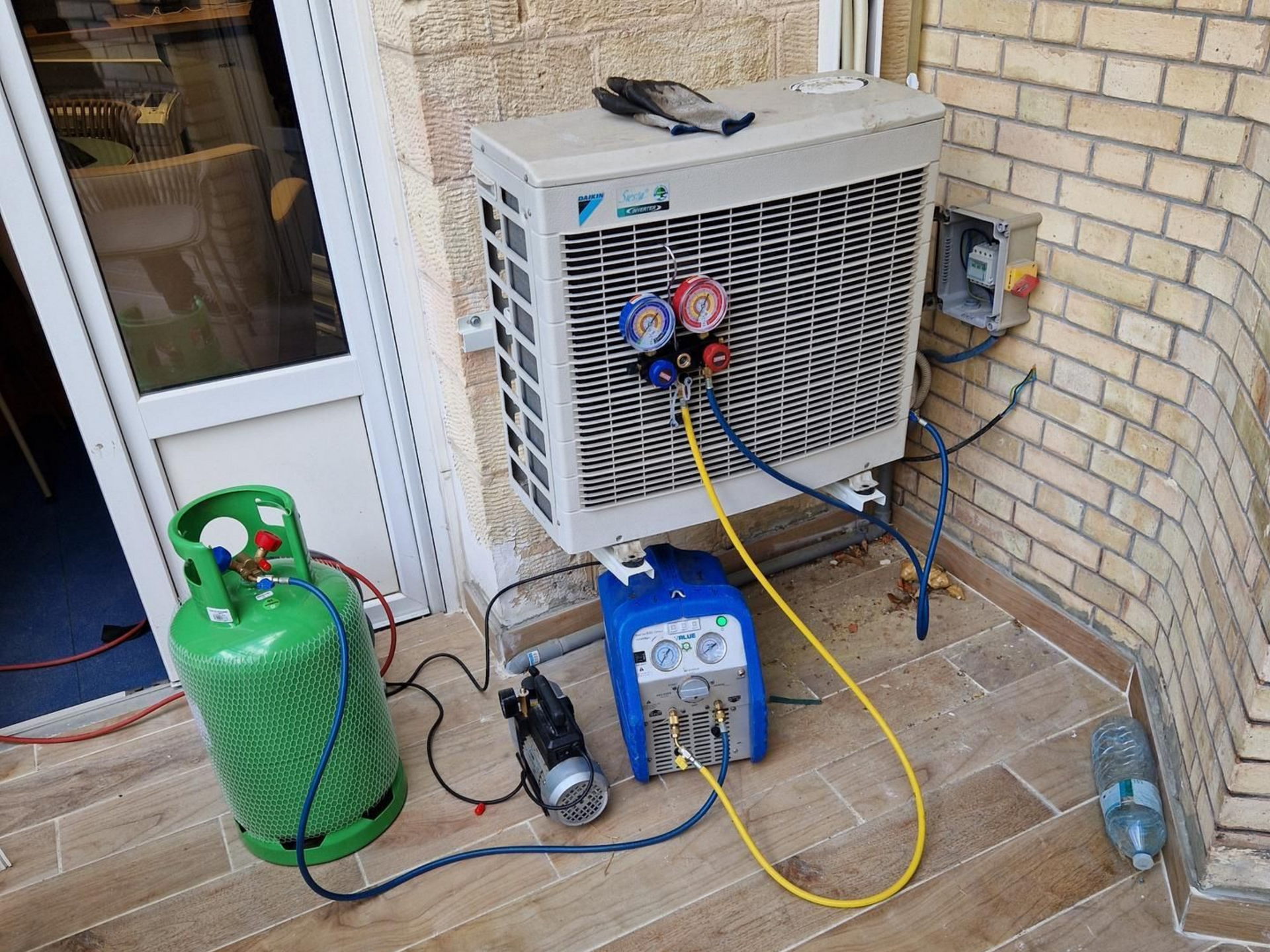Unité de climatisation en cours d'entretien à l'extérieur, équipements connectés : réservoir de réfrigérant, pompe à vide, manomètre, sur une terrasse carrelée.