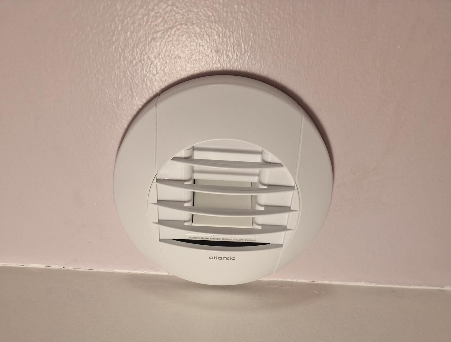 Grille d'aération circulaire blanche dans un plafond rose pâle au-dessus d'un mur blanc.