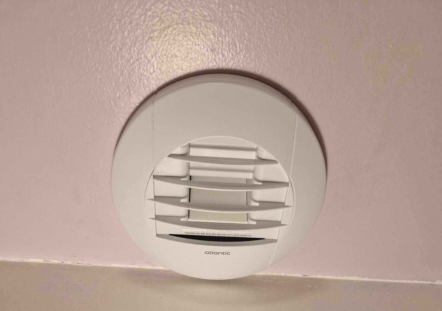 Grille d'aération blanche au plafond rose, au-dessus d'un mur blanc.