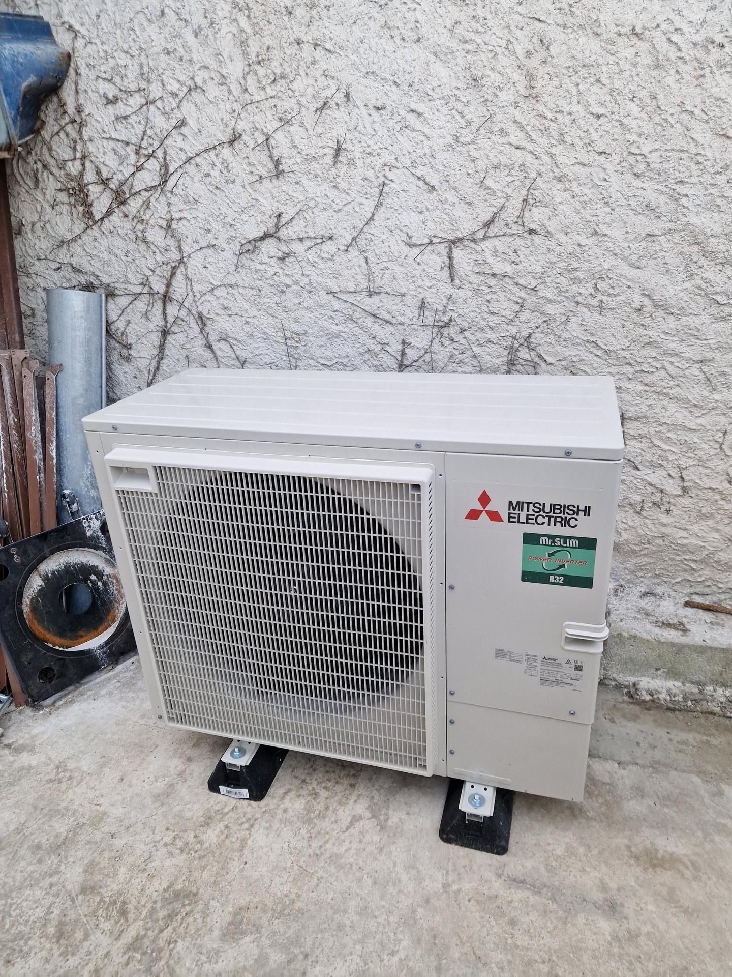 Unité extérieure de pompe à chaleur Mitsubishi sur supports noirs. Contre un mur texturé de couleur claire.
