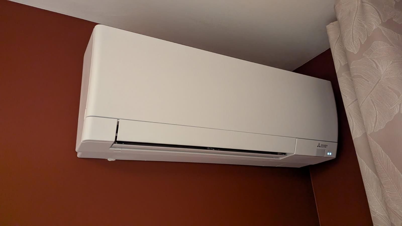 Climatiseur mural blanc sur un mur marron, près d'un plafond et d'un rideau blancs.