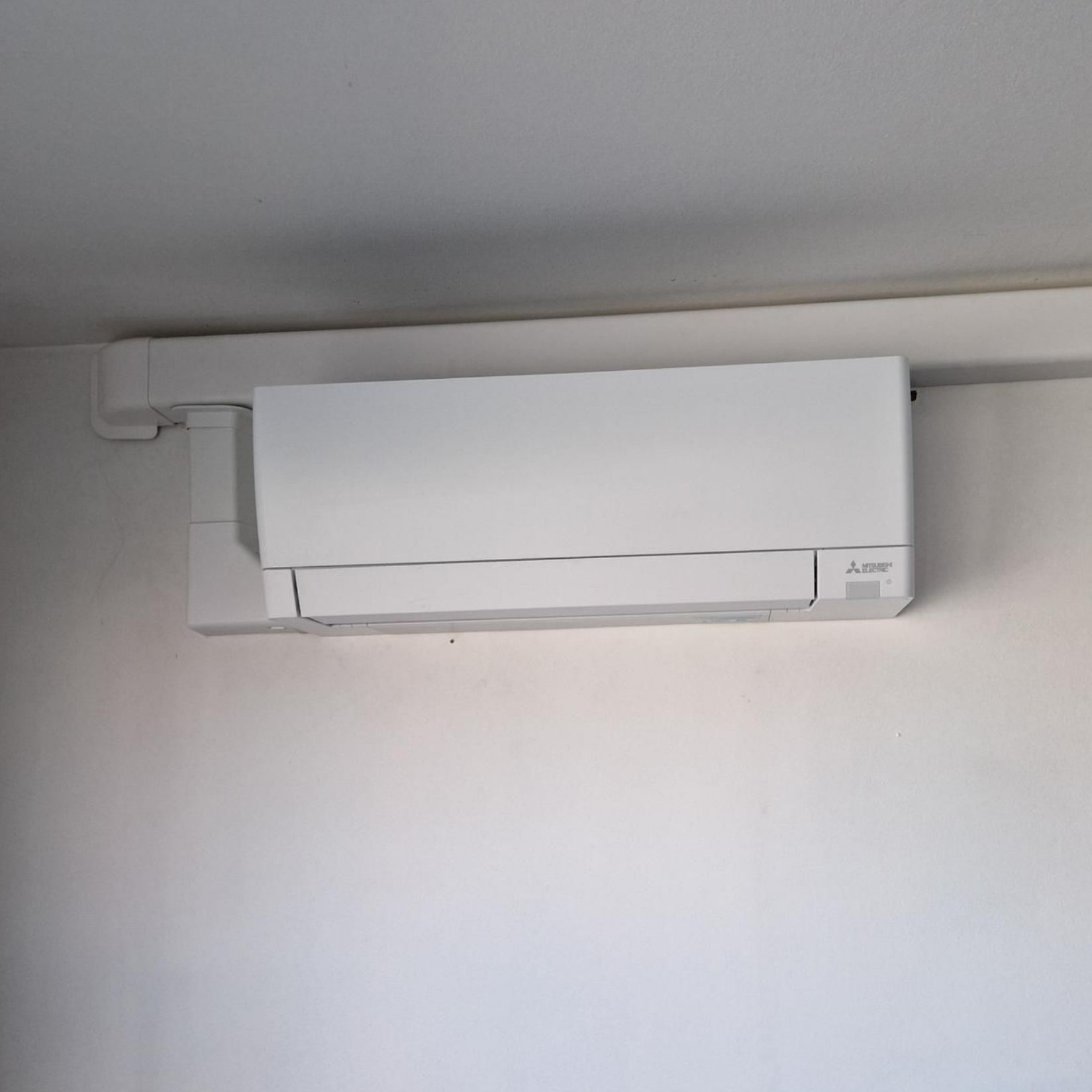 Unité de climatisation blanche fixée sur un mur blanc sous une goulotte de câbles blanche.