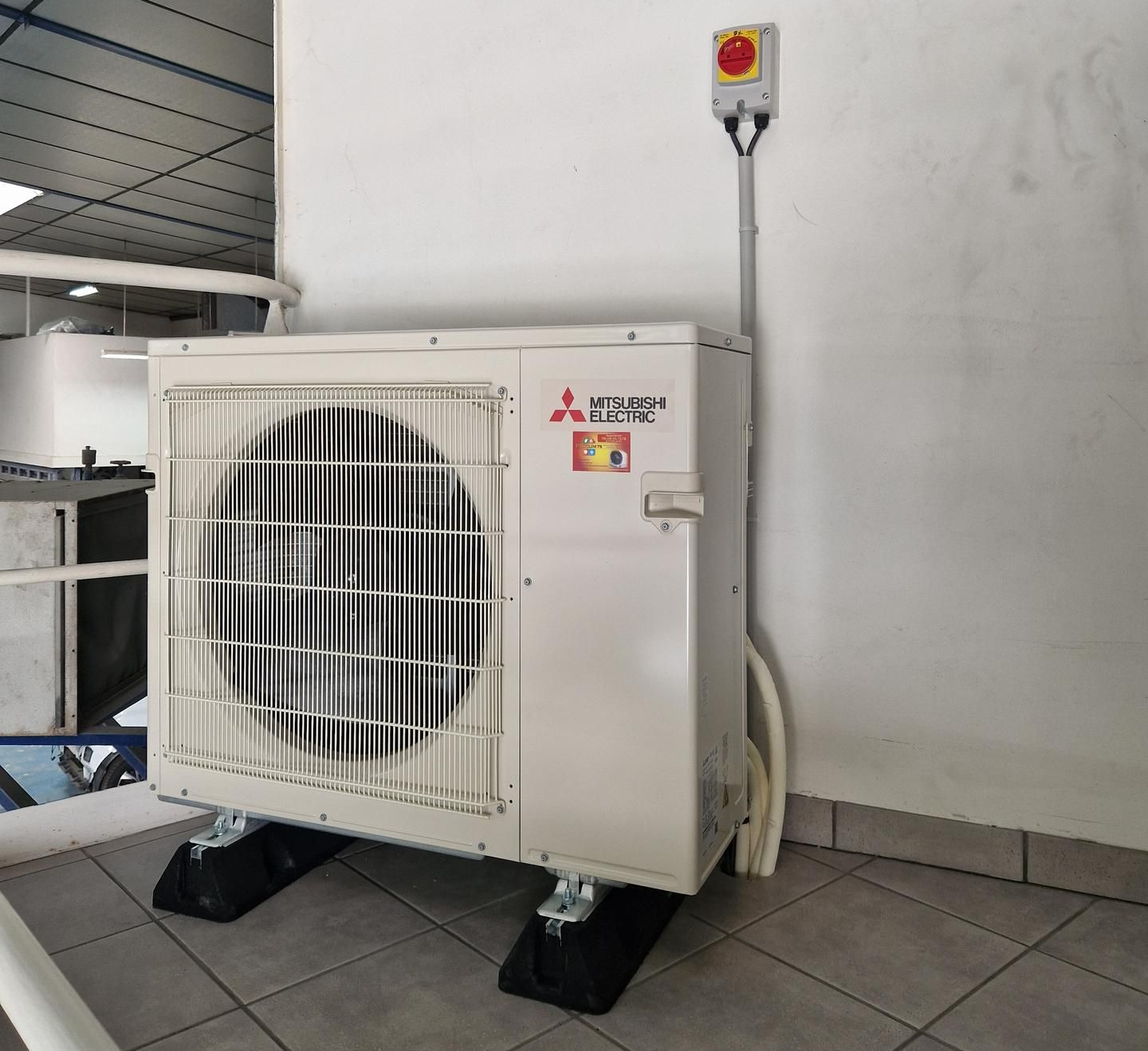 Unité de climatisation sur blocs noirs contre un mur blanc avec bouton d'arrêt d'urgence rouge.