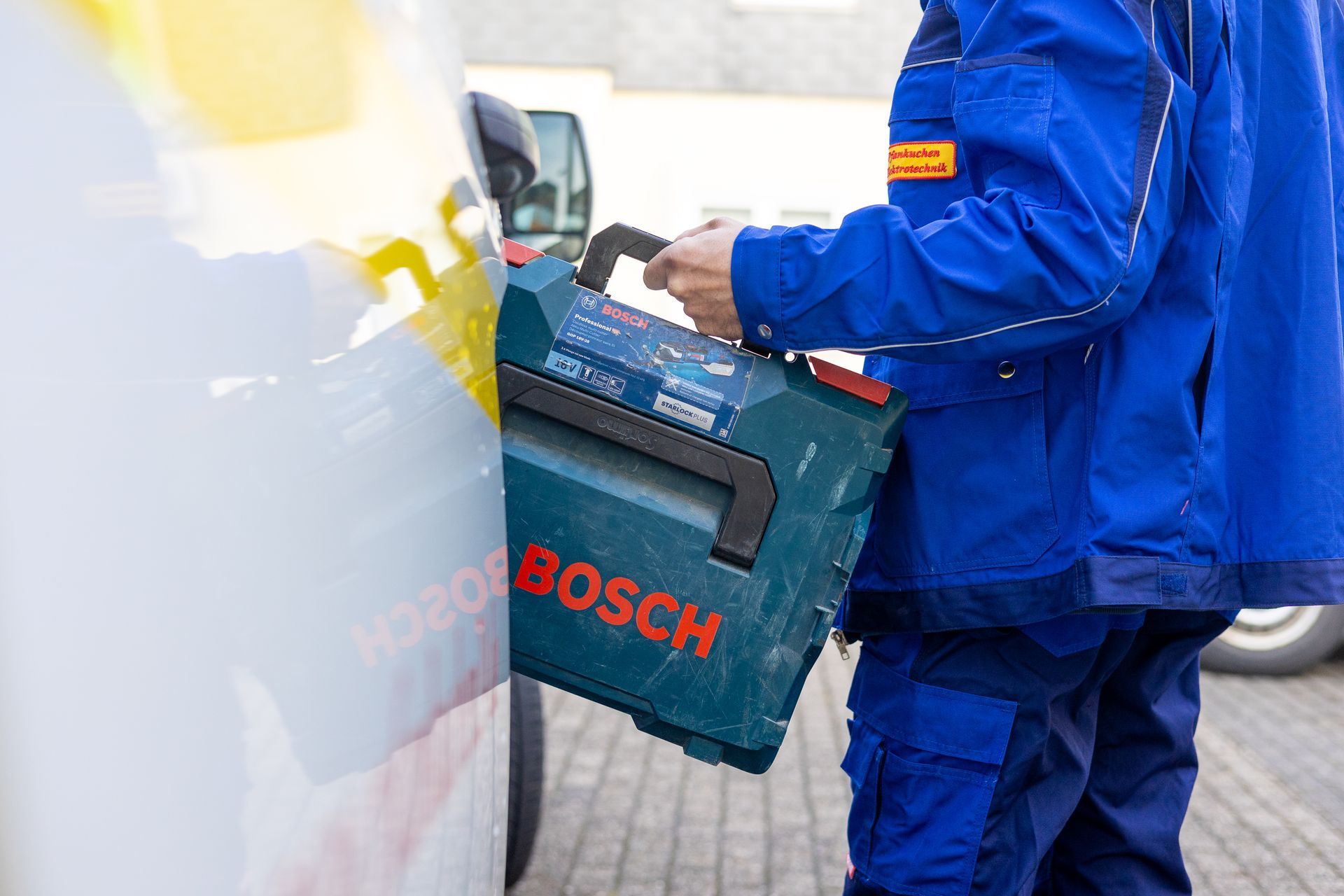 Ein Elektriker in einer blauen Jacke trägt einen Bosch-Werkzeugkasten