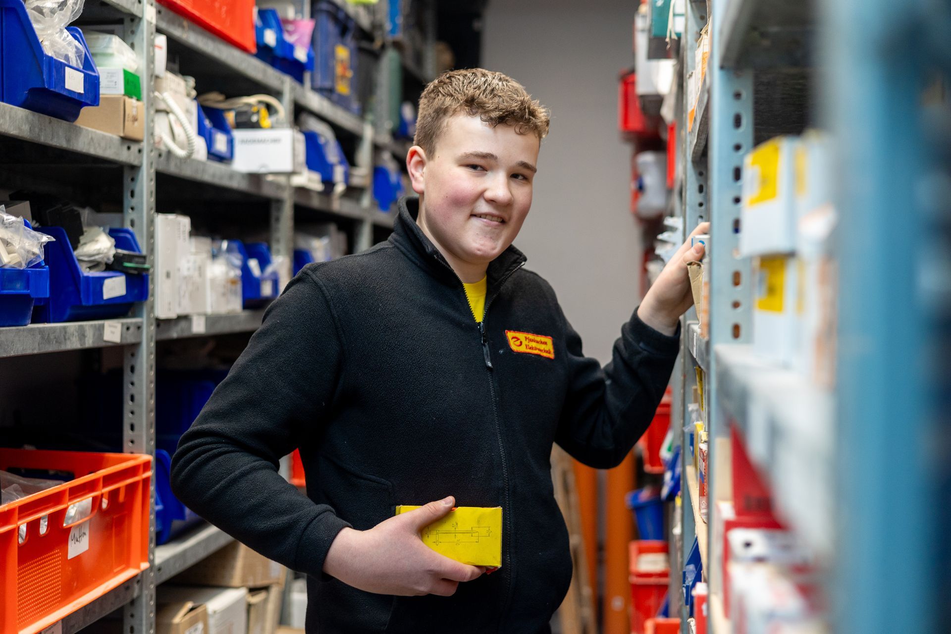 Ein junger Elektriker-Azubi steht in einem Lagerhaus und hält eine gelbe Kiste in der Hand