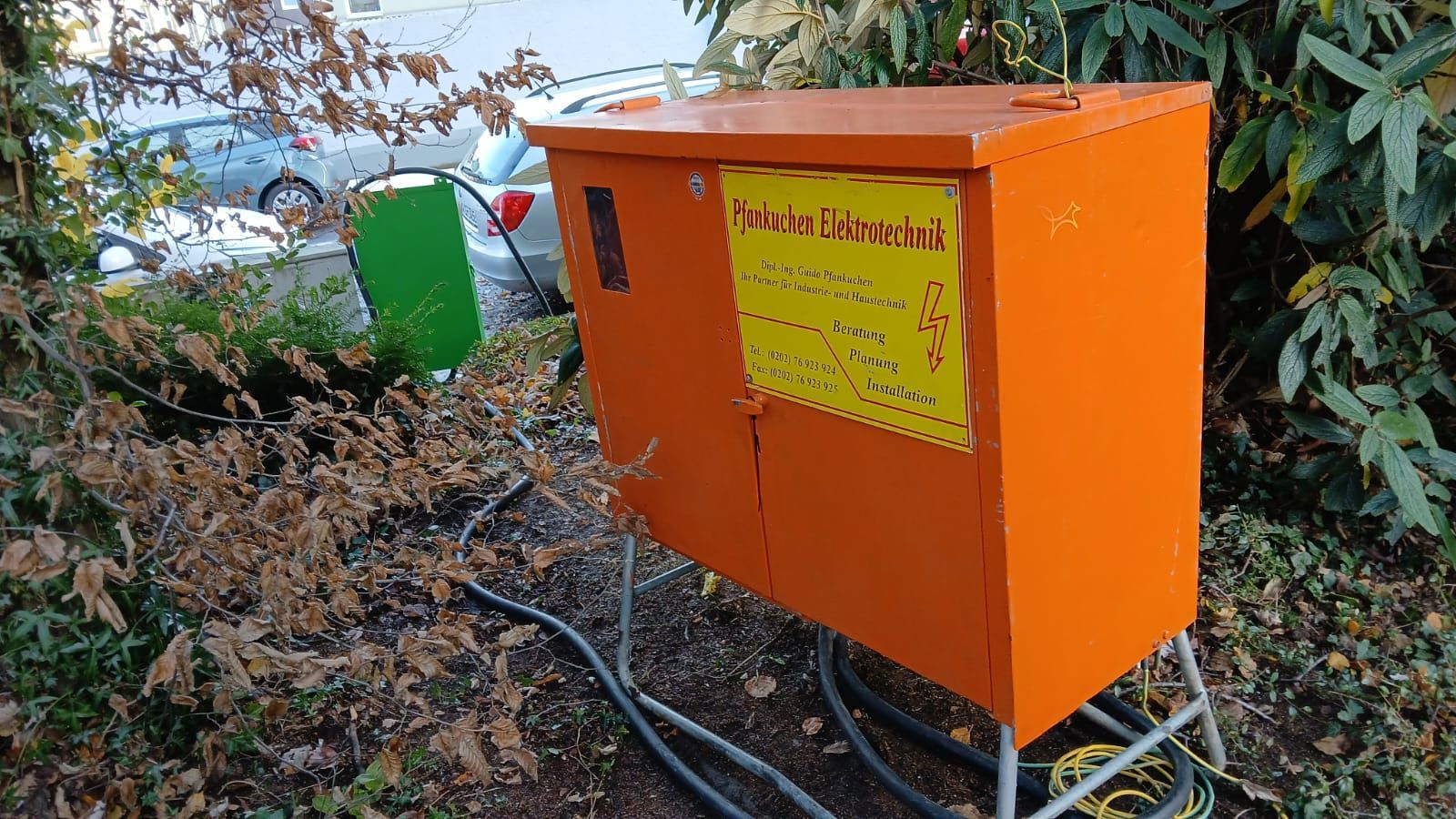 In einem Busch neben einer Baustelle steht ein orangefarbener Baustromkasten