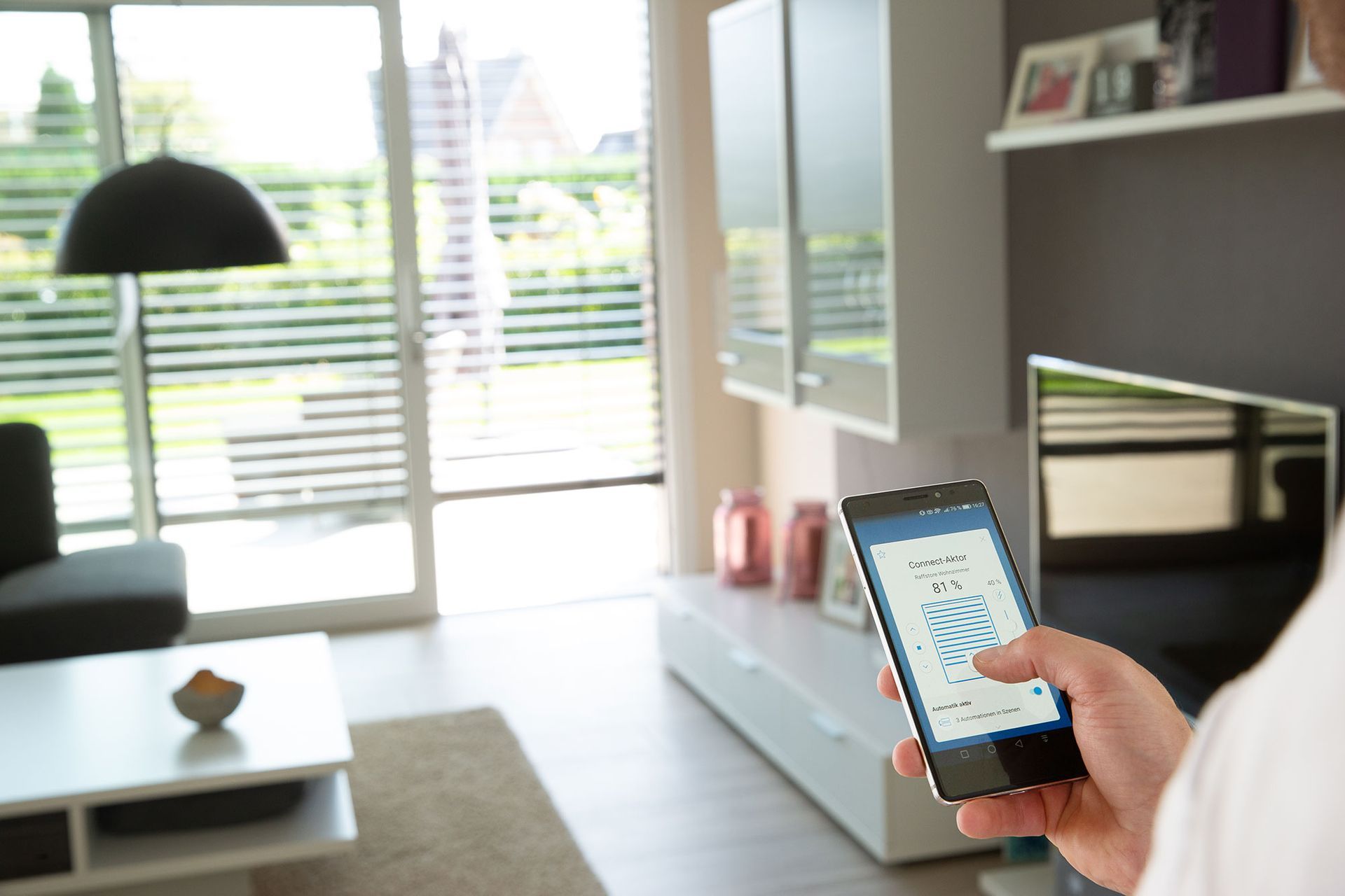 Eine Person sitzt auf einem Sofa und hält ein Tablet mit einer Smart-Home-App in der Hand