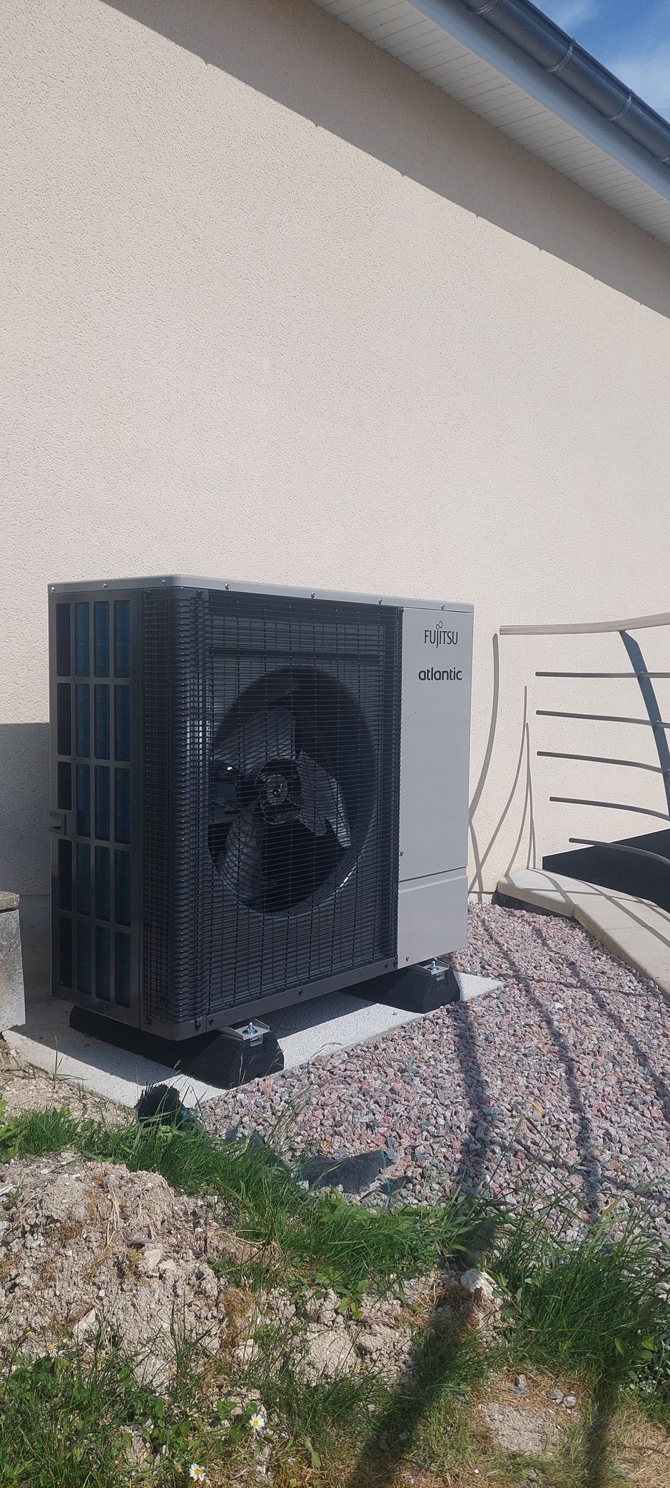 Unité de pompe à chaleur Fujitsu fixée sur un mur blanc, entourée de plantes et de briques.