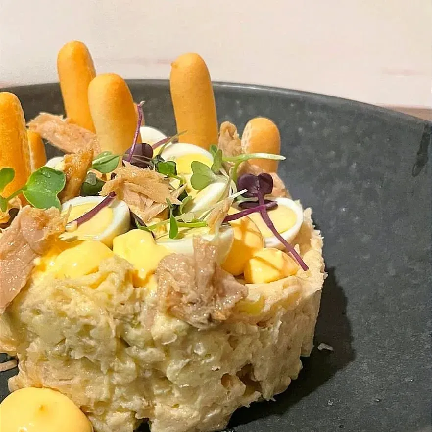 Ensalada de papa cubierta con atún, huevos de codorniz, palitos de pan y microvegetales en un plato negro.