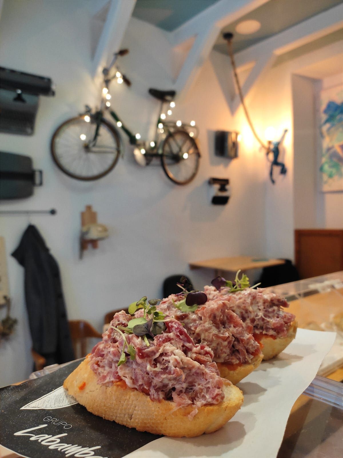 Tres sándwiches abiertos cubiertos con carne y brotes sobre una barra con una decoración de bicicleta de fondo.