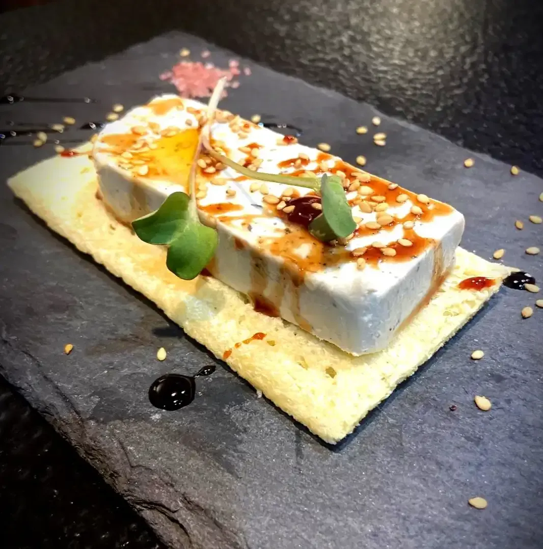Ladrillo de queso blanco con glaseado y semillas de sésamo sobre una galleta, servido en un plato de pizarra.