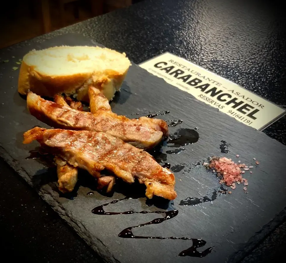 Rebanadas de carne a la parrilla con pan, sal y salsa sobre una pizarra negra. Letrero de restaurante al fondo.