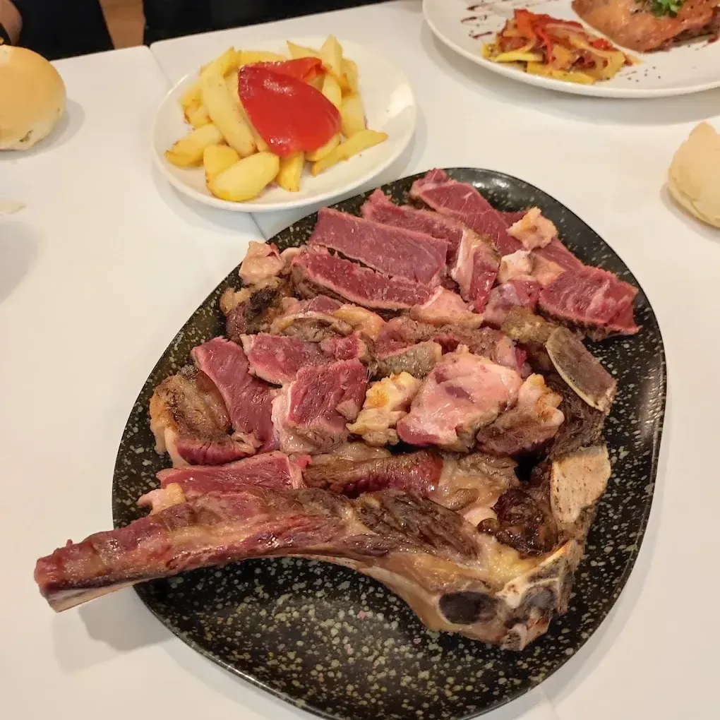 Bistec a la parrilla en un plato oscuro con papas fritas, pimiento rojo y guarniciones sobre un mantel blanco.