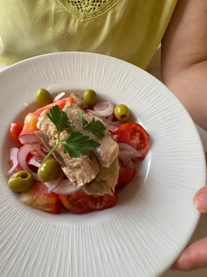 Un plato blanco contiene una ensalada: atún, tomates, cebolla roja, aceitunas y perejil.