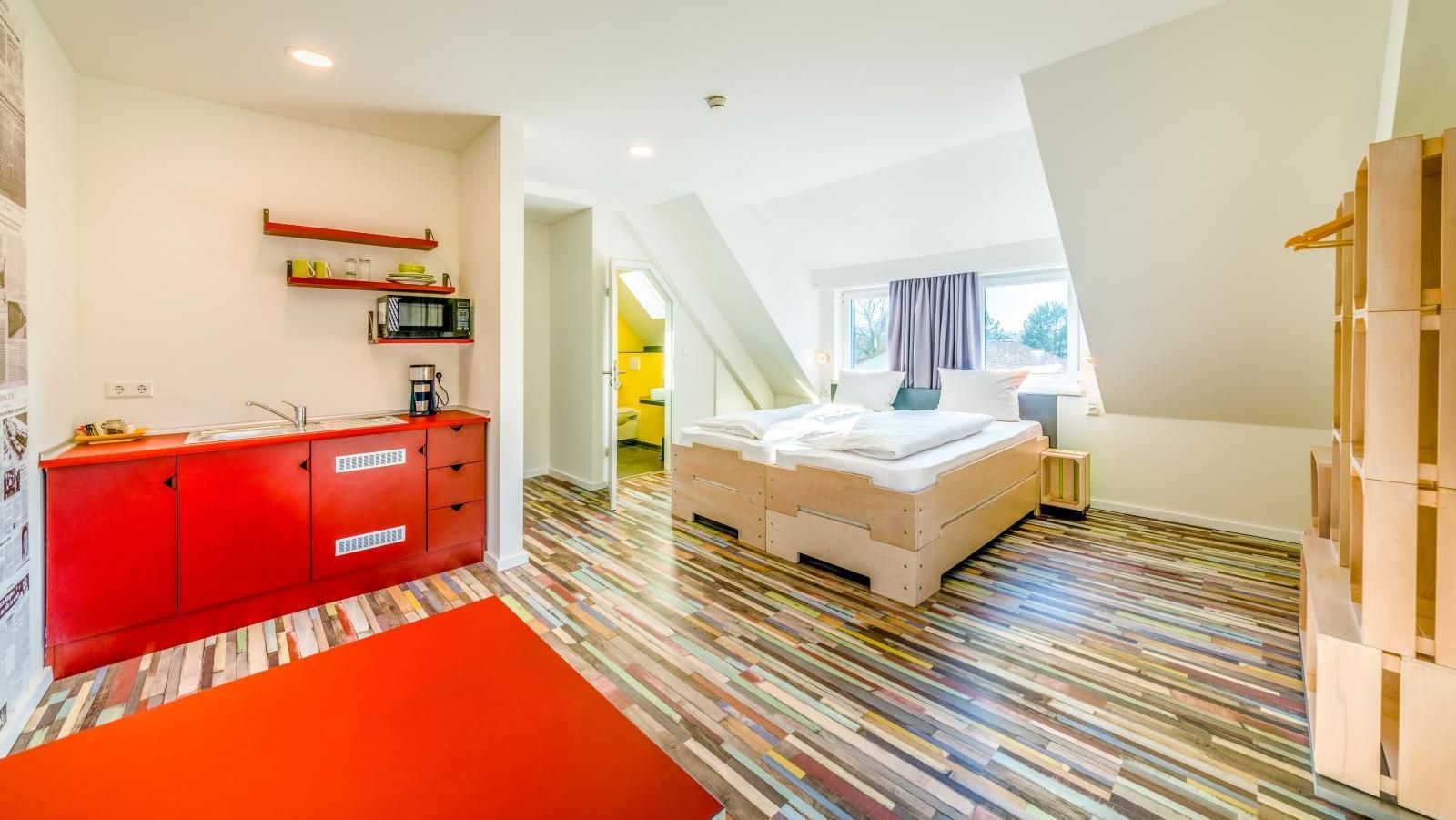 Chambre d'hôtel confortable avec cuisine rouge, lit sous un plafond mansardé, parquet et entrée ouverte.