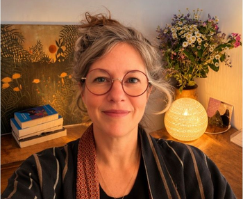 Une femme à lunettes sourit dans un bureau chaleureux à domicile, entourée de livres, de fleurs et d'une lampe qui brille derrière elle.
