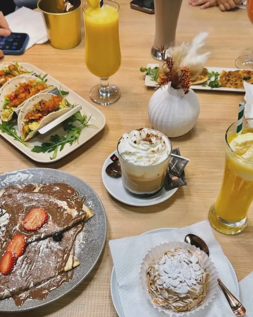 Mesa repleta de tacos, panqueques, pasteles y bebidas de naranja en un ambiente de restaurante informal.