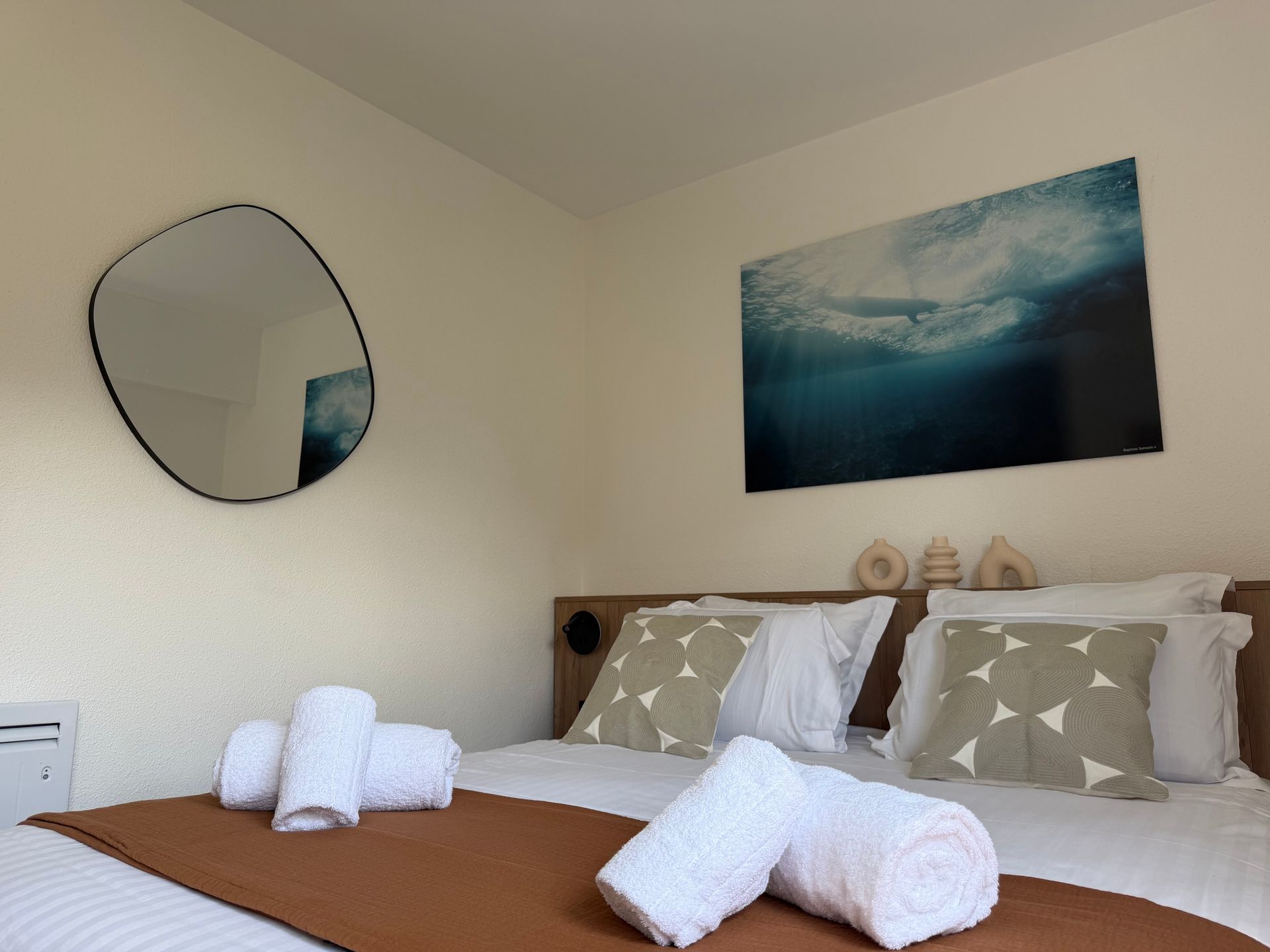 Chambre soignée avec linge de lit blanc, miroir rond mural et tableau représentant l'océan bleu au-dessus du lit.